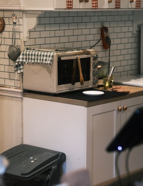 A close-up view of a domestic kitchen counter displaying a small, white oven with a stainless steel handle, situated on a wooden surface. The oven’s exterior appears slightly greasy, with a fabric c